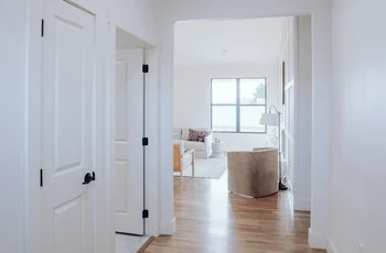 Heller Flur mit Holzboden führt in modernes Wohnzimmer mit großen Fenstern und Sitzmöbeln.